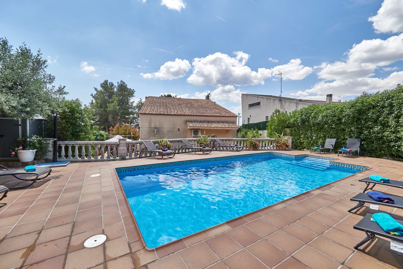 Piscina azul rodeada de jardines y una casa al fondo bajo un cielo soleado.