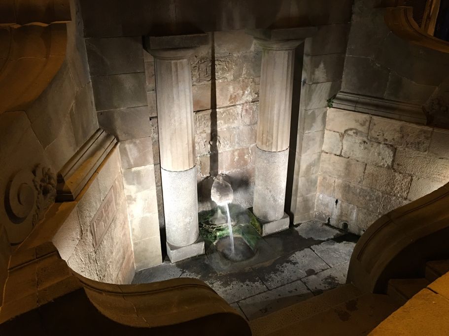 Fuente iluminada al anochecer, rodeada de columnas y muros de piedra.