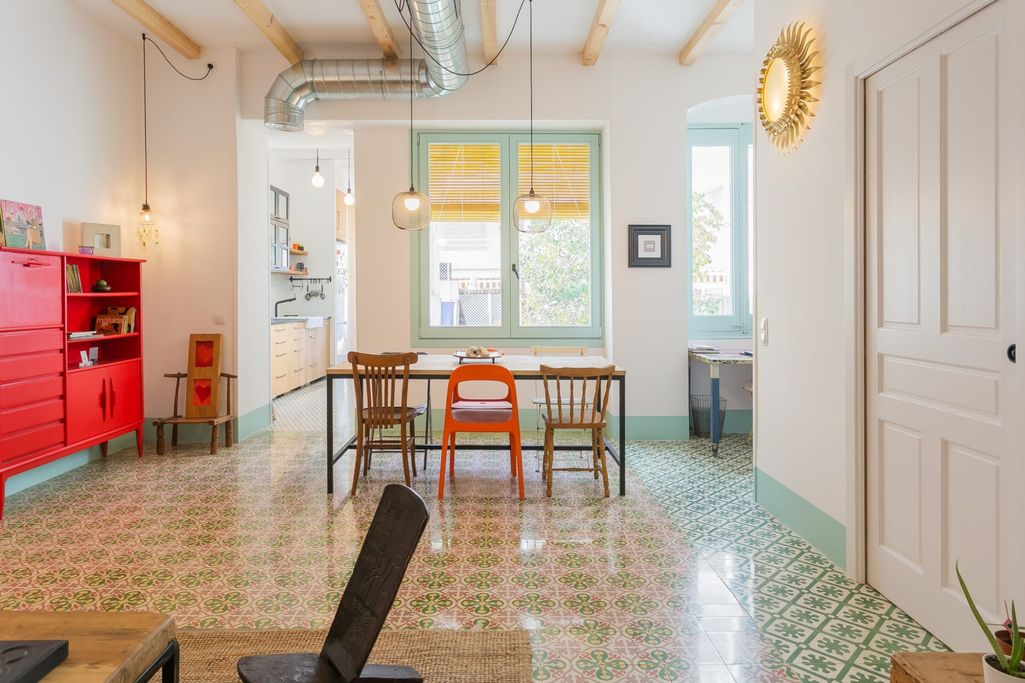 Interior de cocina con mesa de madera, sillas de colores y paredes claras. Suelo de baldosas.