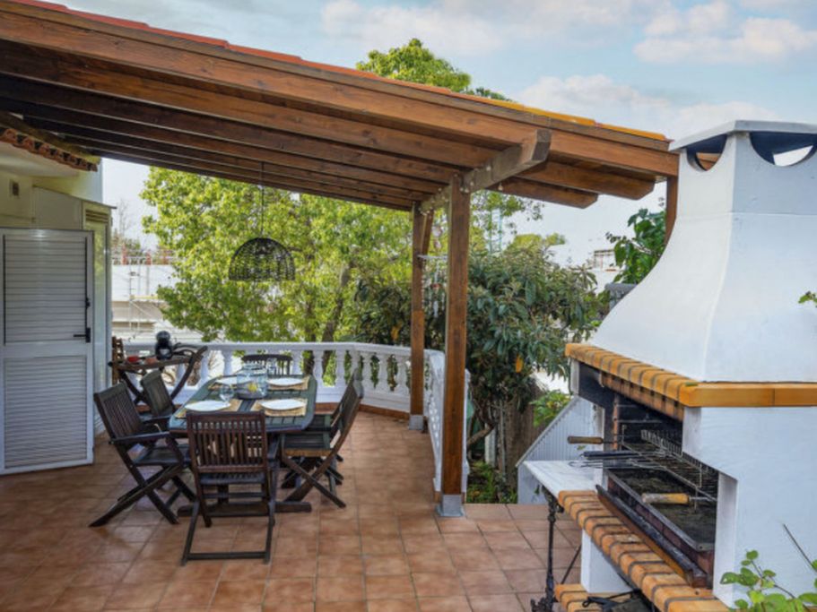 Terraza con mesa, sillas y una parrilla bajo una pérgola rodeada de árboles.